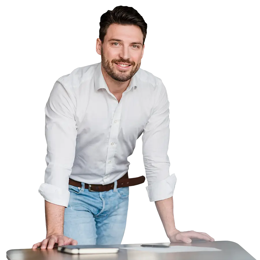 Porträtaufnahme eines Mannes mit dunklen Haaren und Bart in weißem Hemd, braunem Gürtel und heller Jeans, der sich an einen Tisch lehnt und lächelt.